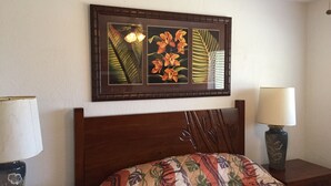 Une chambre à coucher avec une tête de lit en bois, un lit recouvert d’une couvre-lit à motifs floraux, une table de chevet avec une lampe et, au mur, un tableau encadré représentant des fleurs et des feuilles.