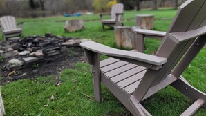 Primer plano de una silla Adirondack gris con estructura metálica, ubicada en una zona cubierta de césped con un brasero al fondo.