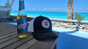 A bottle of Kalik beer and a black cap with a logo on a wooden table by a pool with a view of the ocean and palm trees. - Modern vacation home with private yard, steps to the beach and centrally located (Mt Thompson)
