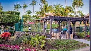 A covered outdoor dining area with a grill, surrounded by palm trees and a stone wall.