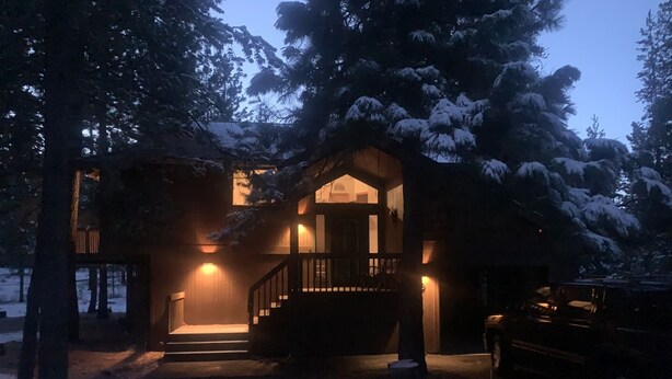 Ein mit Schnee bedecktes Haus mit einer Holztreppe, die zu einer Veranda führt, umgeben von Bäumen.