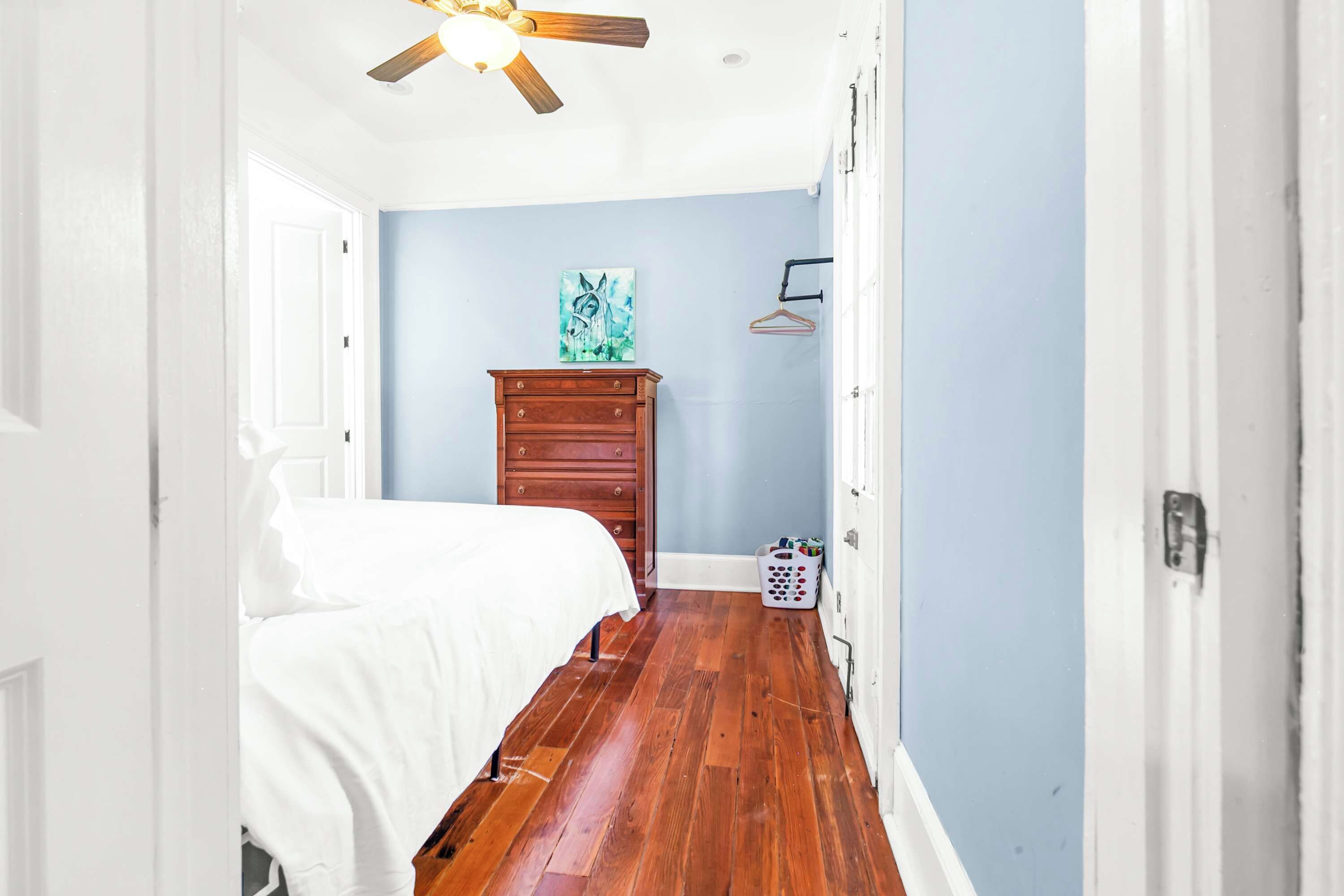 A narrow hallway with a wooden floor, a bed with white bedding, a dresser, a painting on the wall, and a laundry basket.