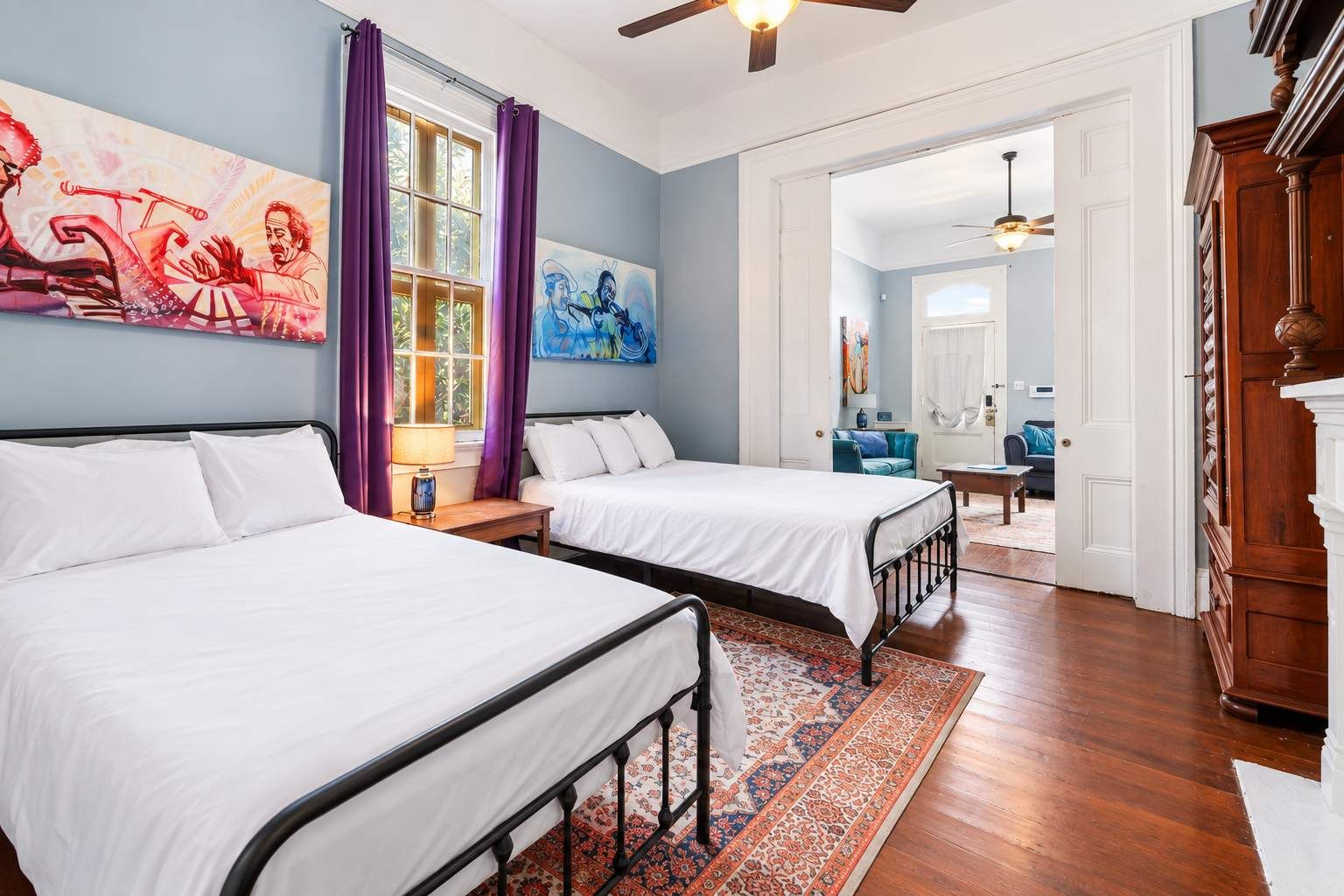 Une chambre avec deux lits, une fenêtre avec des rideaux violets, un ventilateur de plafond et un tableau accroché au mur.