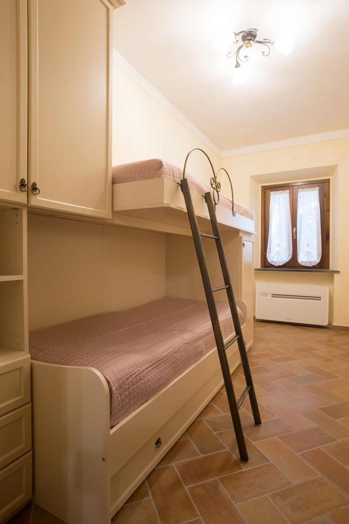A bunk bed room with a ladder, a window with curtains, and a ceiling light fixture.