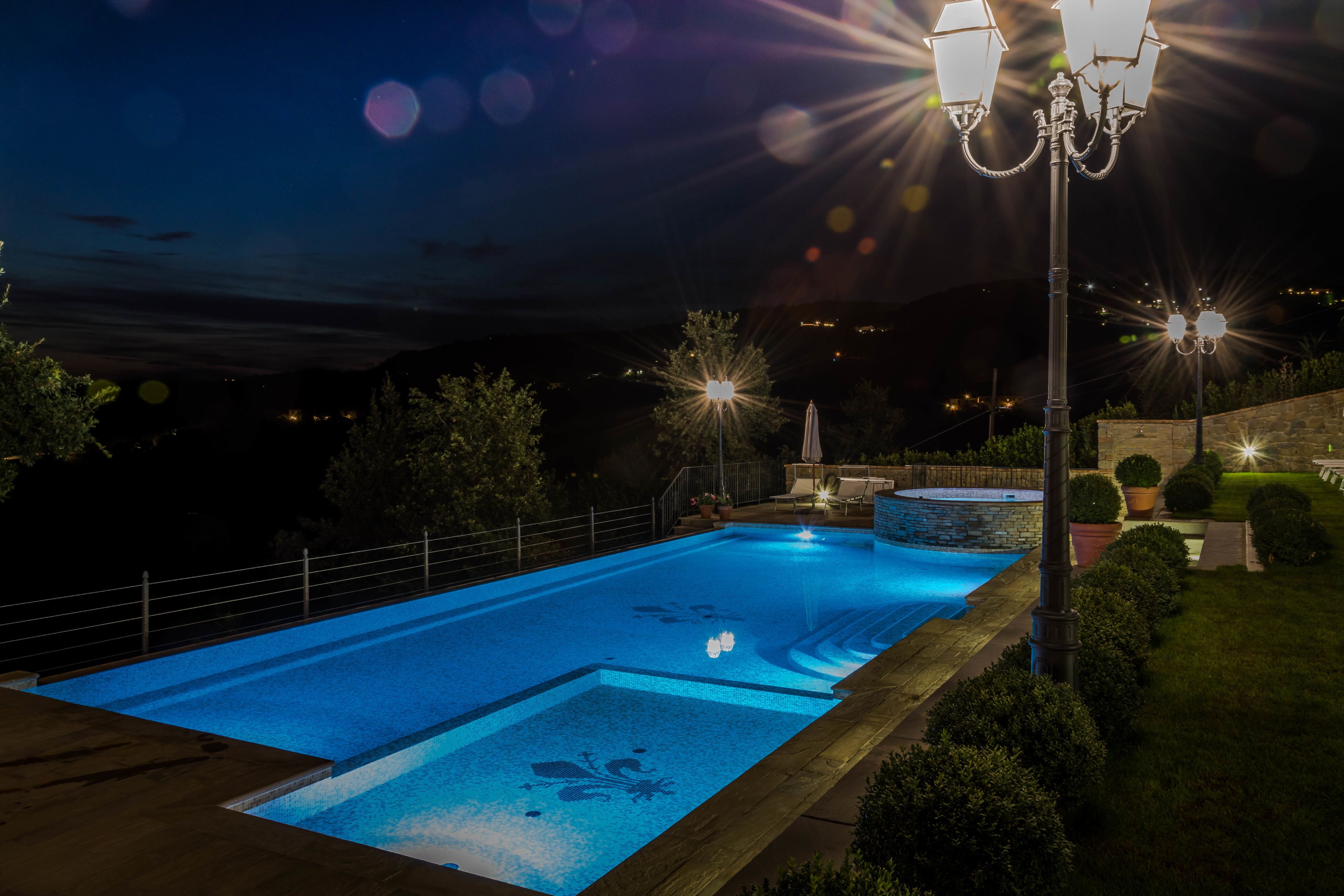 A swimming pool at night with surrounding greenery and a stone wall.