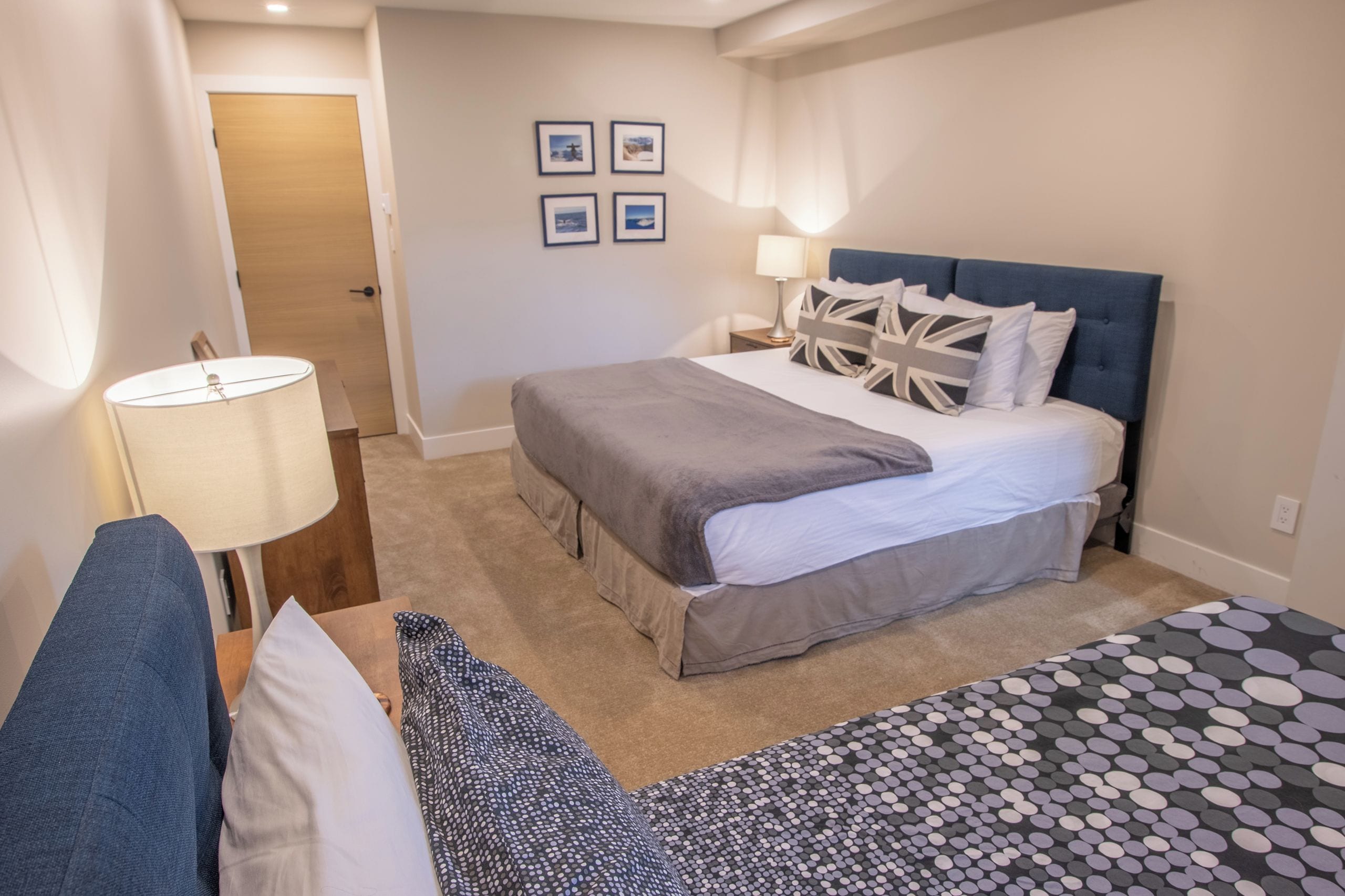 A hotel room with a large bed, a bedside table with a lamp, a chair, and a small table. There are four framed pictures on the wall.