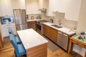 A modern kitchen with stainless steel appliances, wooden cabinets, a white countertop, and a dining area with blue upholstered chairs.