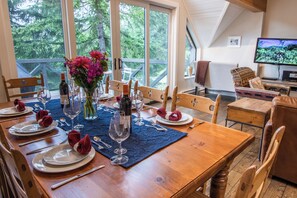 A dining area with a wooden table set for six, surrounded by chairs. A large window offers a view of trees. - Pinnacle Ridge 36 - Ski in Ski out, Private Hot Tub, Wood Fireplace (Whistler)