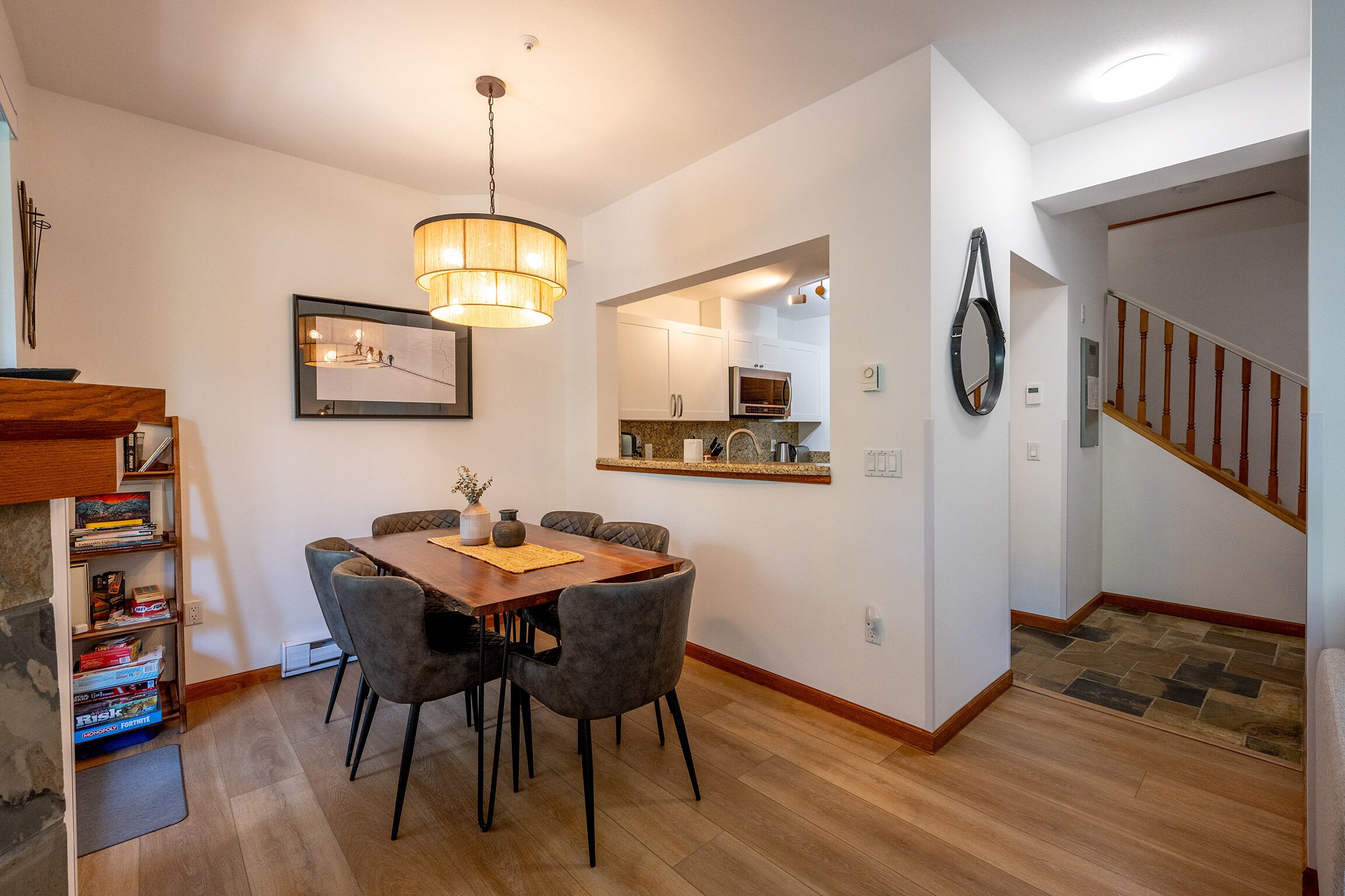 A dining area with a wooden table, grey upholstered chairs, a hanging light fixture, and a kitchen visible through an archway.