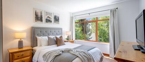 A bedroom with a large bed, a nightstand with a lamp, a TV on a wooden stand, and a window with a view of trees.
