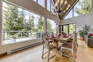 A dining area with a large table and chairs, large windows with a view of trees, and a chandelier.