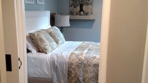 Ein Schlafzimmer mit einem Bett, einem Nachttisch, einer Lampe und einem an der Wand befestigten Regal mit einem Baumgemälde.