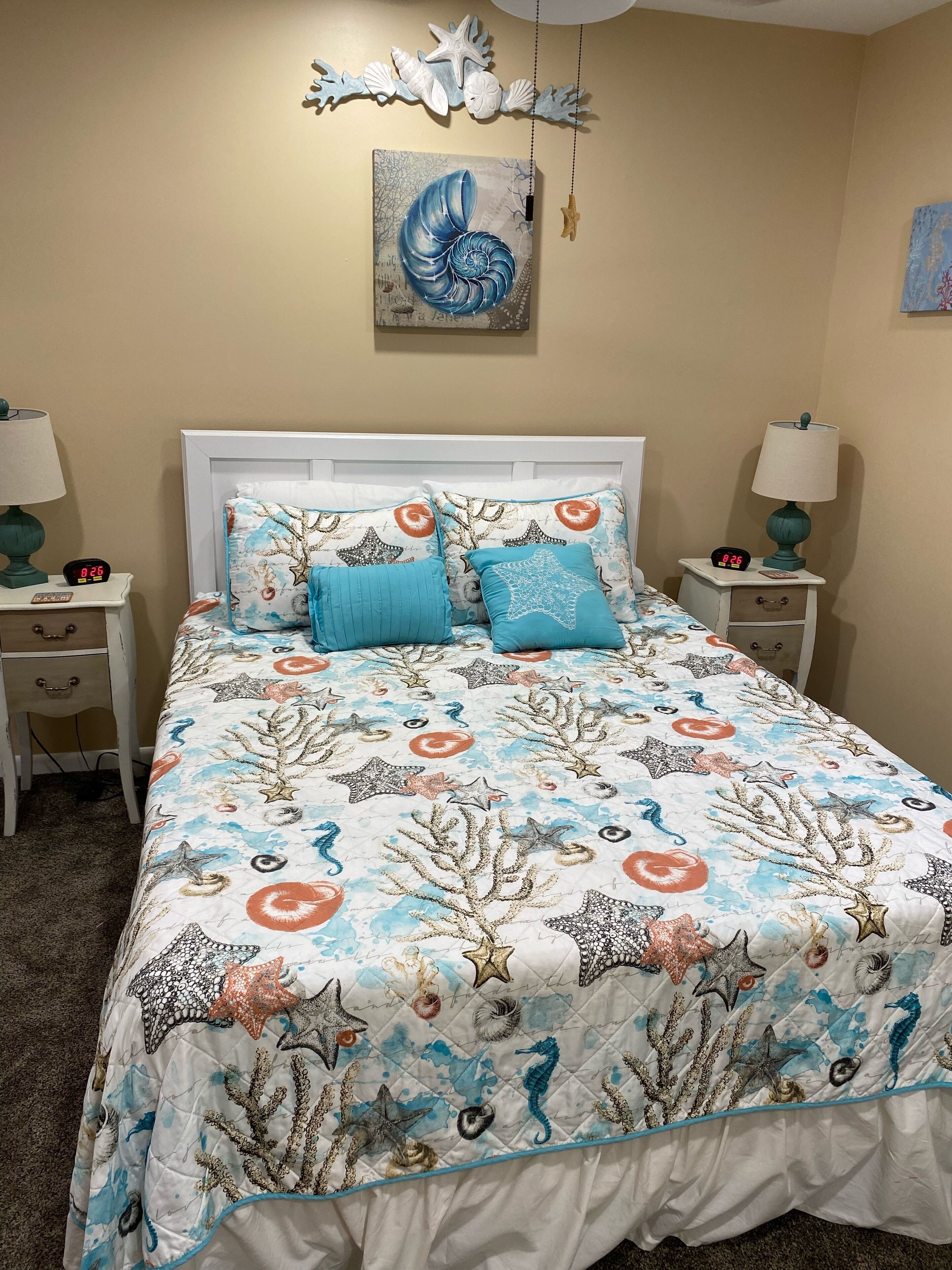 A bedroom with a bed featuring a nautical-themed bedspread, two bedside tables with lamps, a wall clock, and a framed artwork above the bed.