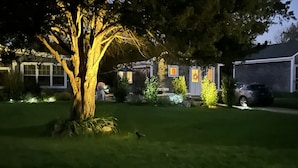 A residential area at night with a well-lit house, a driveway, and a tree in the foreground.