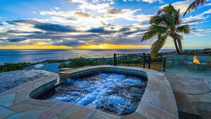 A hot tub with a curved edge, surrounded by stone tiles, overlooking a body of water and palm trees under a sunset sky.