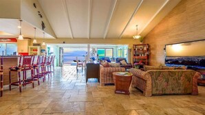 A spacious living area with a large window offering a view of the ocean, wicker furniture, and a flat-screen TV mounted on the wall.