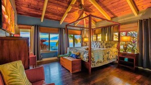 A four-poster bed with a view of the ocean, wooden flooring, and a ceiling fan.