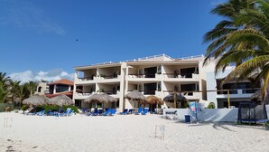 Un hôtel situé en bord de mer, doté de nombreux balcons, de parasols en chaume et de chaises longues bleues.