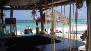 Un bar situé en bord de mer, doté d’un grand comptoir noir, d’un toit de chaume et offrant une vue sur l’océan.