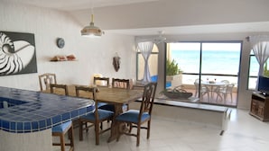 Un espace repas avec une table et des chaises en bois, un comptoir de bar aux carreaux bleus et une vue sur l’océan grâce à de grandes fenêtres.