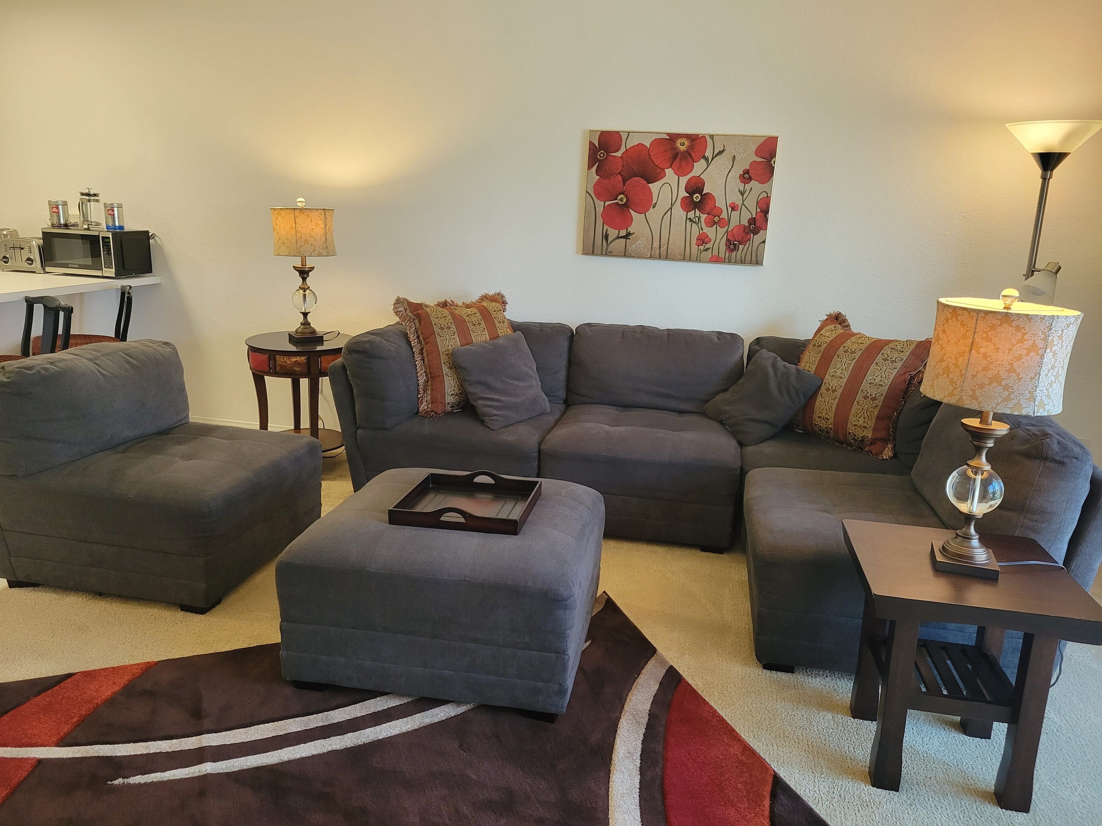 Ein Wohnzimmer mit einer grauen Eckcouch, einem kleinen Hocker, einem Couchtisch, einem Beistelltisch mit Lampe, einem Gemälde von roten Blumen und einer Küchenzeile mit Mikrowelle und Kaffeemaschine.