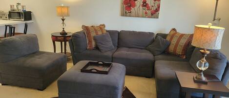 Ein Wohnzimmer mit einer grauen Eckcouch, einem kleinen Hocker, einem Couchtisch, einem Beistelltisch mit Lampe, einem Gemälde von roten Blumen und einer Küchenzeile mit Mikrowelle und Kaffeemaschine.