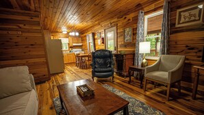A cozy cabin interior with a wooden floor, a sofa, a dining table, and a kitchen area. - Mountaineer Cabin 6 - Hot Tub & Gas Fireplace close to New River Gorge (Hico)