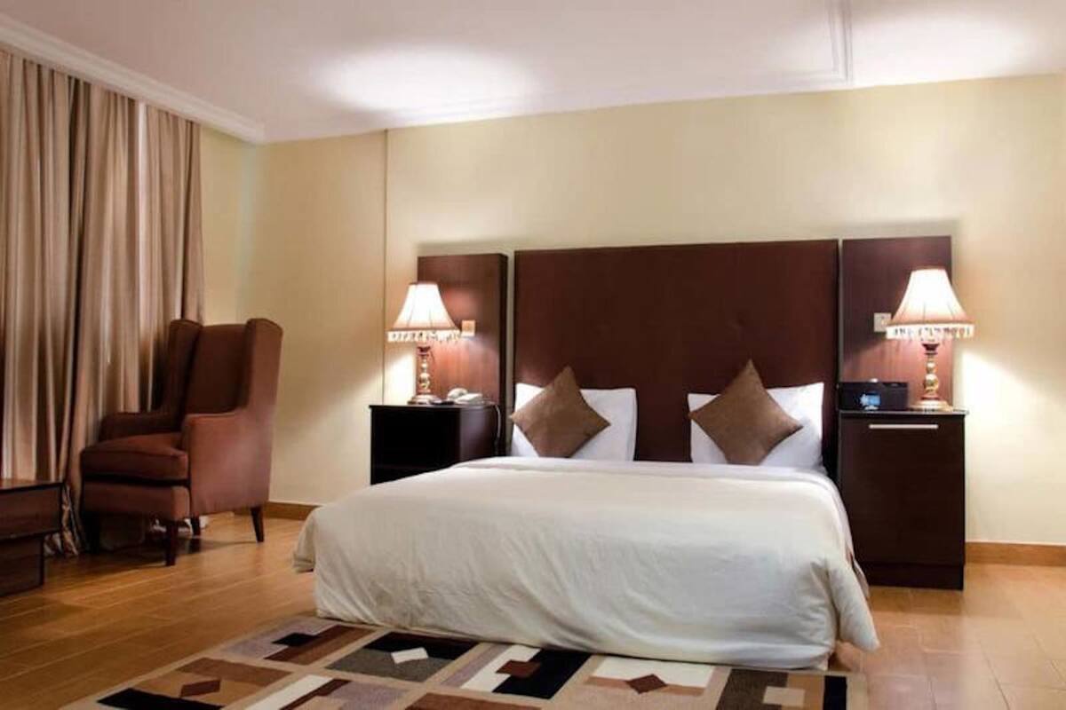 Quarto de hotel com uma cama grande, duas mesas de cabeceira com luminárias, uma poltrona marrom e um tapete estampado.