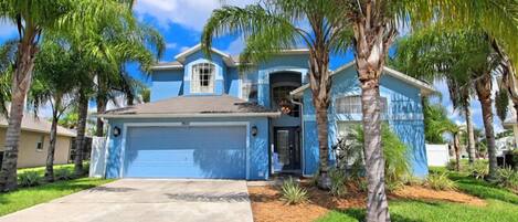 A blue two-story house with a garage, surrounded by palm trees and a well-maintained lawn.