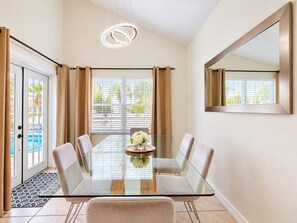 A modern dining area with a glass table, six chairs, a chandelier, and a view of a pool through large windows. - 10 minutes to Disney! Two Pools & Spa! No Carpet! Themed Rooms @The Nemo House (Kissimmee)