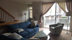 A living room with a blue sofa, patterned cushions, a glass coffee table, and a patterned armchair. A staircase is visible in the background.