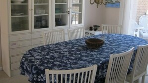 A dining area with a round table covered in a blue floral tablecloth, surrounded by white chairs. A cabinet with glass doors is visible in the background.