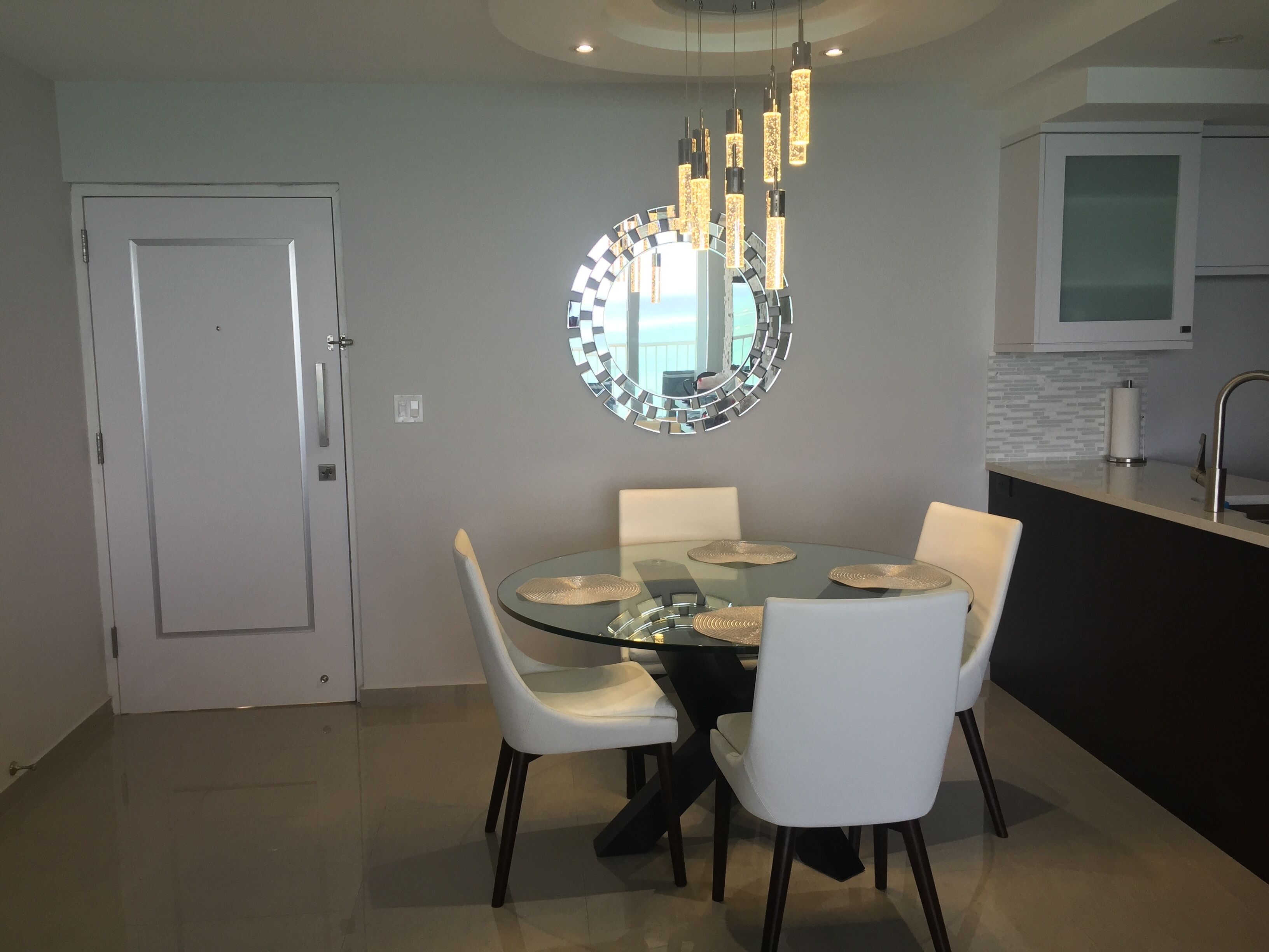 A modern dining area with a glass table, white chairs, and a unique circular mirror above.
