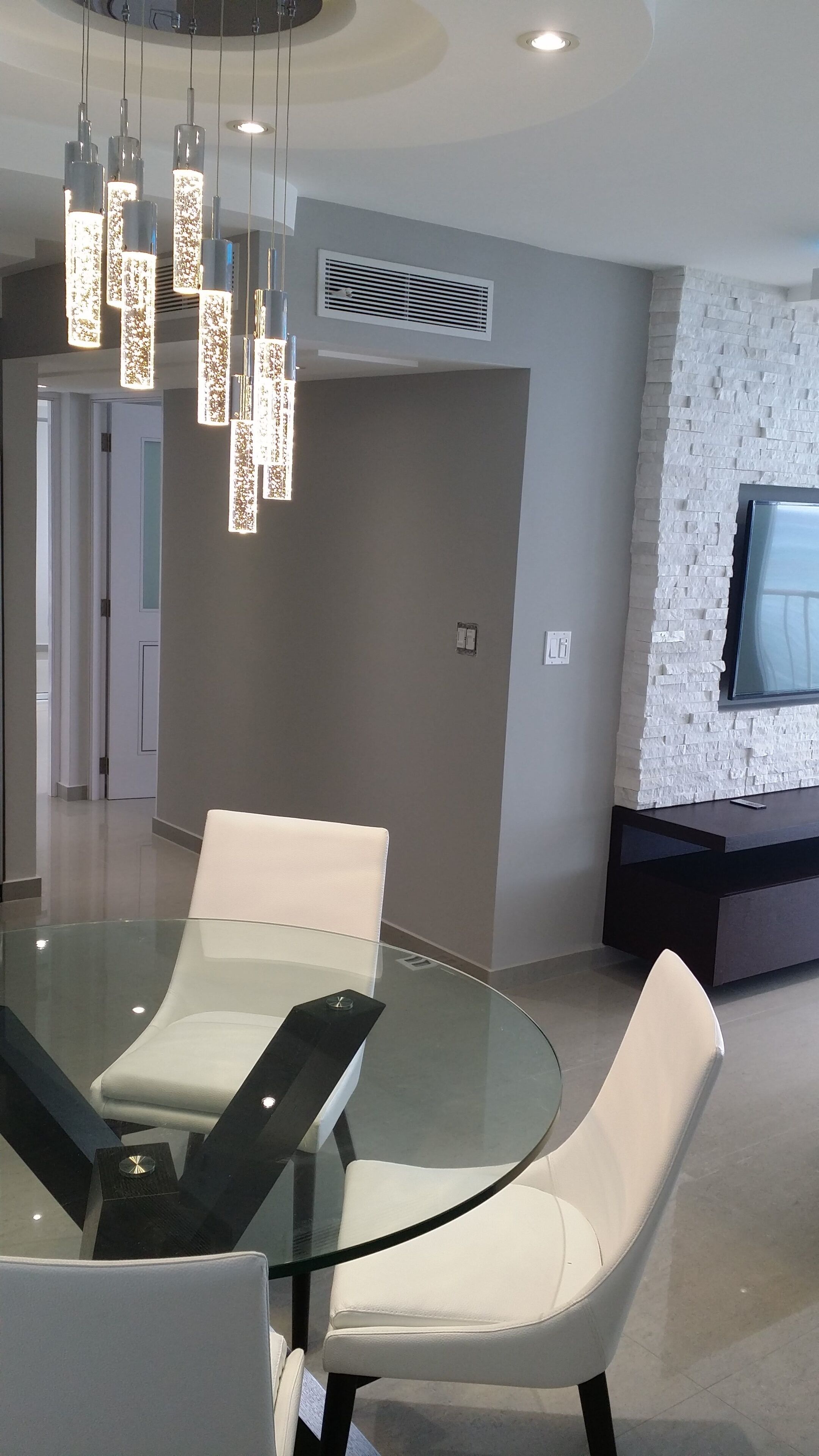 A modern dining area with a glass table and white chairs, a ceiling light fixture, and a TV mounted on a brick-patterned wall.