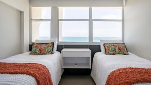 A bedroom with two beds, a window with a view of the sea, and a white bedside table. - Marbella del Caribe Condo - Amazing Oceanfront View (Carolina)