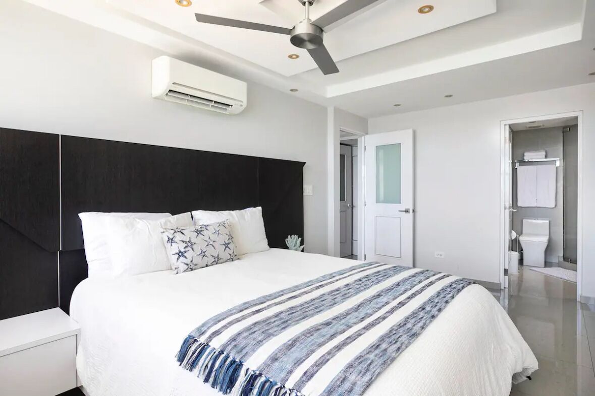 A modern bedroom with a large bed, a ceiling fan, an air conditioning unit, and a bathroom with a toilet and sink.