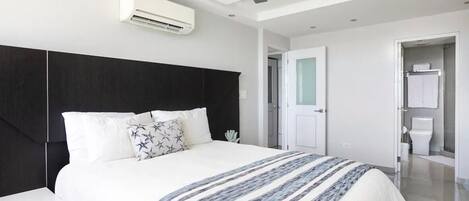 A modern bedroom with a large bed, a ceiling fan, an air conditioning unit, and a bathroom with a toilet and sink.