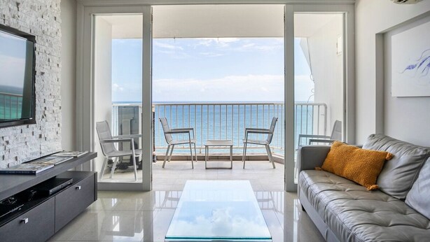 A modern living room with a balcony, a flat-screen TV, a leather sofa, and a glass coffee table. - Marbella del Caribe Condo - Amazing Oceanfront View (Carolina)