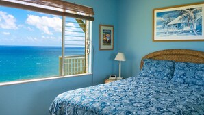 A bedroom with a large window, a bed with a blue patterned bedspread, a framed picture on the wall, and a nightstand with a lamp. - Gorgeous Ocean Views From All Rooms! Corner Unit! Cool AC! Aloha Galore! (Princeville)