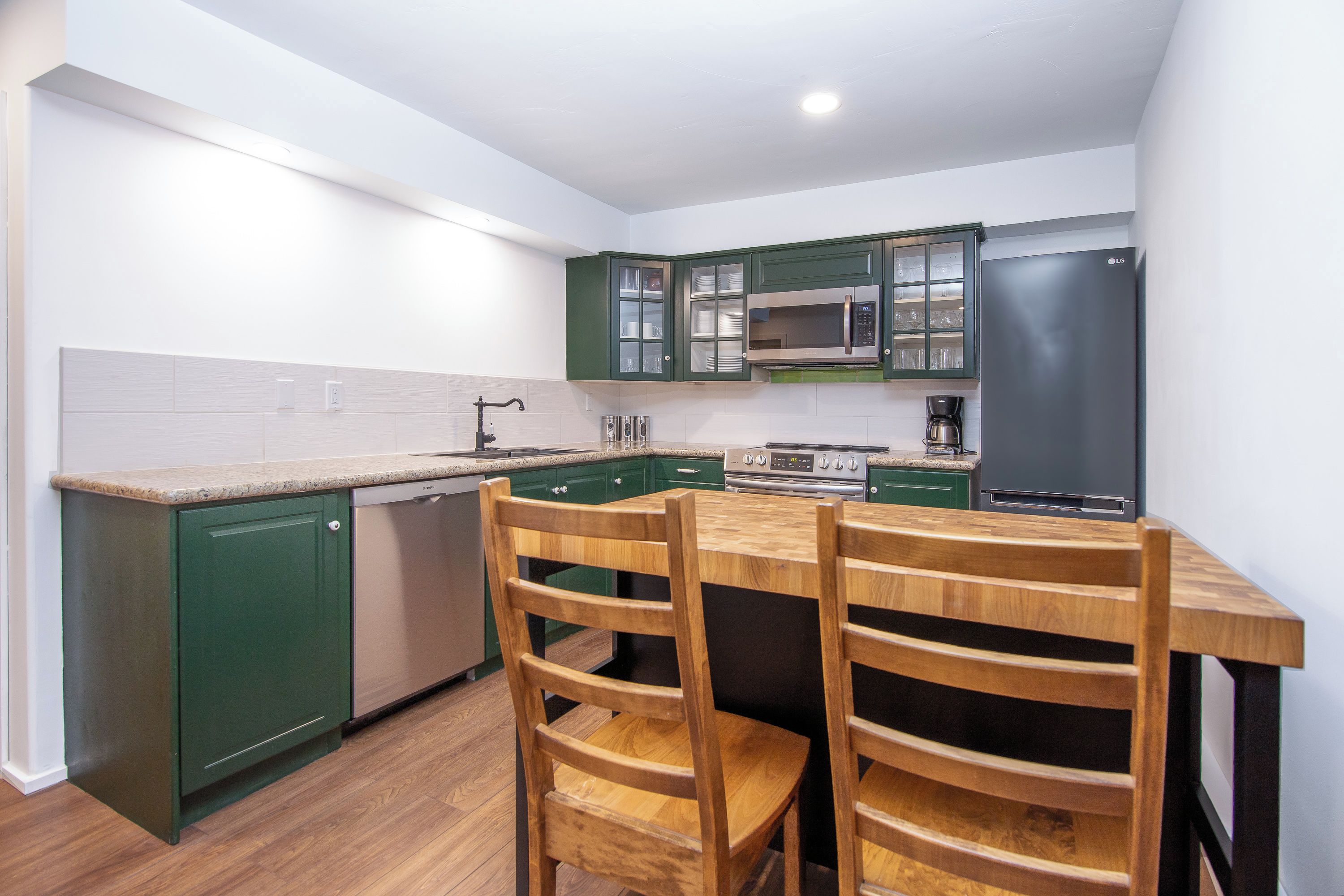 Een kleine keuken met groene kastjes, een magnetron, een koelkast en een houten eettafel met stoelen.