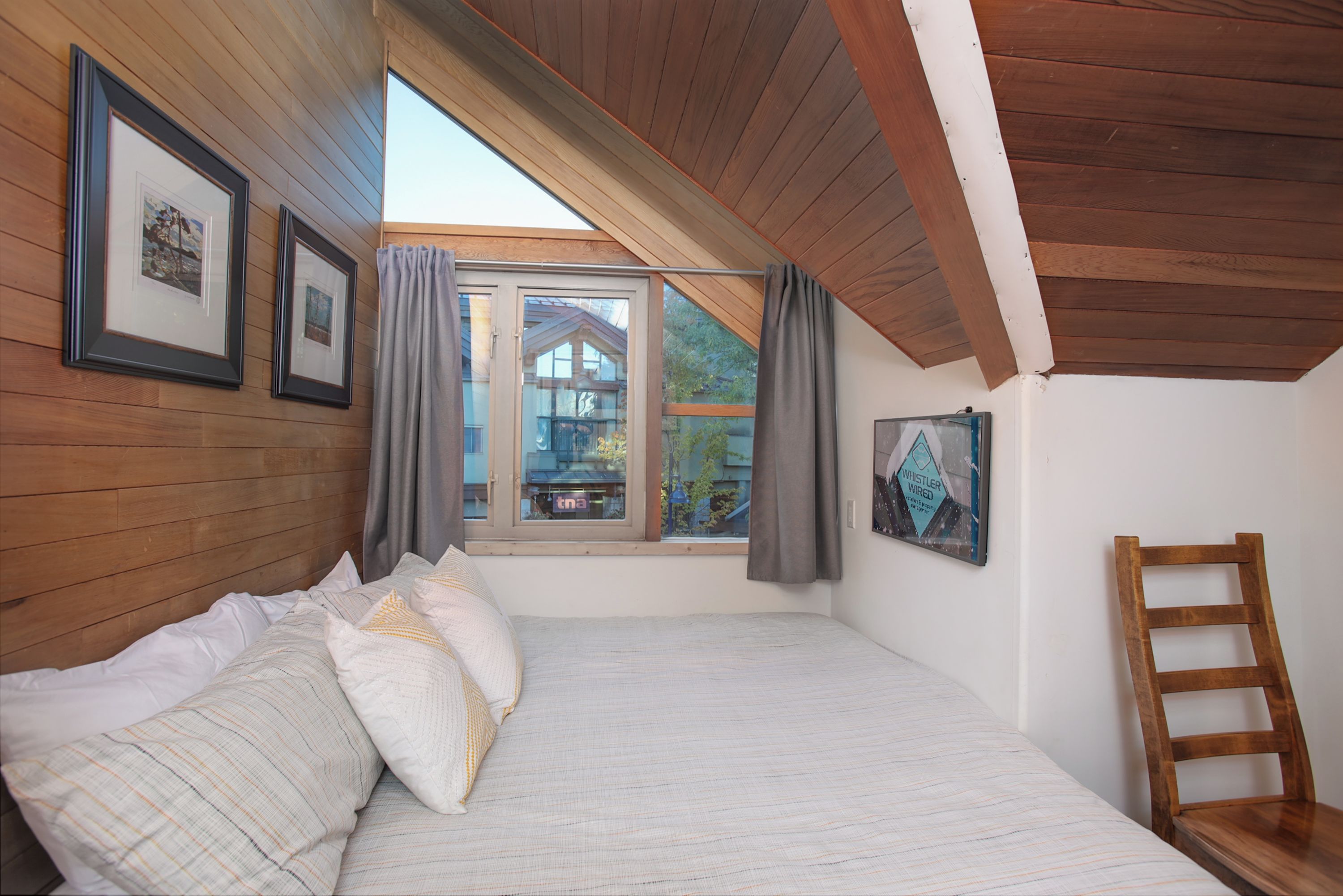 A bedroom with a sloped ceiling, wooden walls, a bed with pillows, a window with curtains, and a chair.