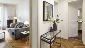 Een moderne woonkamer met een grijze bank, een bijzettafeltje met een lamp en een glazen salontafel. Er hangt een spiegel aan de muur en op een bijzettafeltje staat een vaas met witte bloemen.