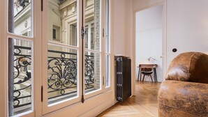 Een kamer met grote ramen, een radiator, een bruine leren fauteuil en een kleine tafel met een lamp.