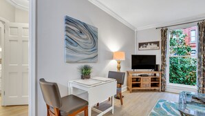 A modern living room with a white desk, a comfortable chair, a TV, and a large window with a view of greenery. - Romantic Condo in City Center- Wright Square Loft C (Savannah)