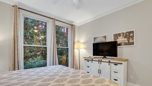 A bedroom with a bed, a TV on a wooden cabinet, a window with curtains, and two framed pictures on the wall. - Romantic Condo in City Center- Wright Square Loft C (Savannah)