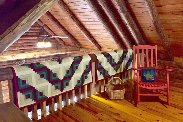 Una habitación cálida en el ático con techo de madera, una manta estampada sobre una barandilla, una silla roja y una cesta en el suelo.