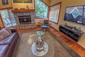 Una sala de estar con chimenea de piedra, sofá de cuero, mesa de centro de vidrio y televisor montado en la pared.