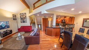 A spacious living area with a red sofa, a glass coffee table, and a dining table with chairs. A kitchen with wooden cabinets and stainless steel appliances is visible in the background. - Snowy Creek 4 - Ski in Ski out, Private Hot Tub, Short Walk to Whistler Village (Whistler)