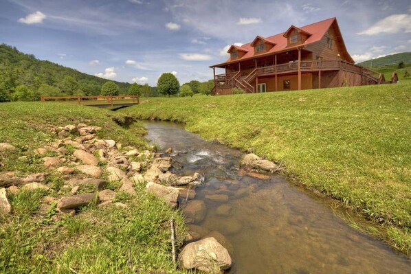 Une maison rustique en bois, avec un toit rouge, un petit ruisseau qui traverse un champ verdoyant et une colline en arrière-plan.
