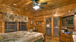 A wooden cabin bedroom with a bed, a ceiling fan, a television, and a bookshelf.